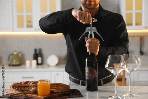 Romantic dinner. Man opening wine bottle with corkscrew at table in kitchen, closeup
