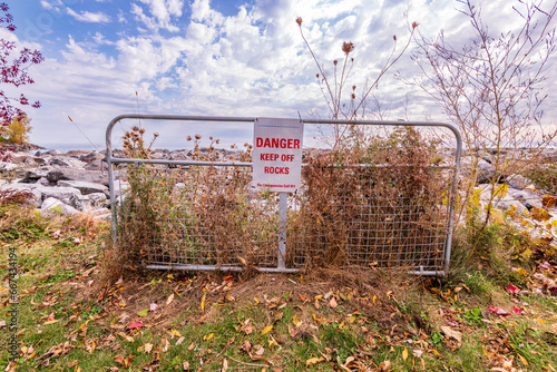 danger keep off the rocks: signage on a portable metal barrier surrounded by weeds with puffy white clouds in background room for text shot in ashbridges bay in fall photo