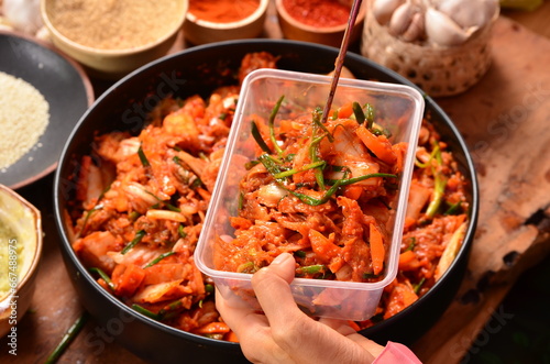  picks up kimchi in a plastic container with chopsticks from a bowl of shredded vegetables, including scallions, cabbage and carrots. There are rows of condiments, including Korean chilies. photo