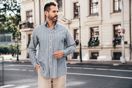 Portrait of handsome confident stylish hipster lambersexual model. Sexy modern man dressed in blue shirt and trousers. Fashion male posing on street background in Europe city at sunset