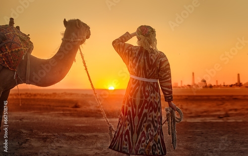 Woman in traditional national clothing leads camel through desert towards ancient city of Khiva at sunset. photo