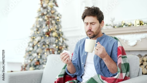 Sick man with a cold or flu is suffering from a cough sitting on sofa at home during winter New Year Christmas Xmas holidays. Male wrapped in blanket wipes his nose with a handkerchief, drinks hot tea photo