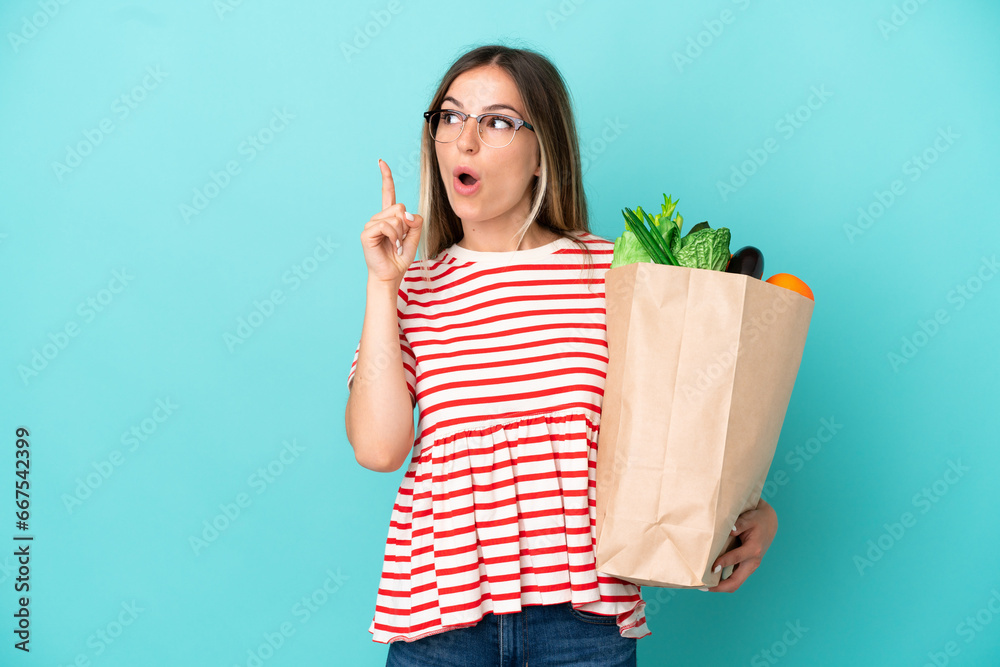 Young woman holding a grocery shopping bag isolated on blue background thinking an idea pointing the finger up Young woman holding a grocery shopping bag isolated on blue background thinking an idea pointing the finger up