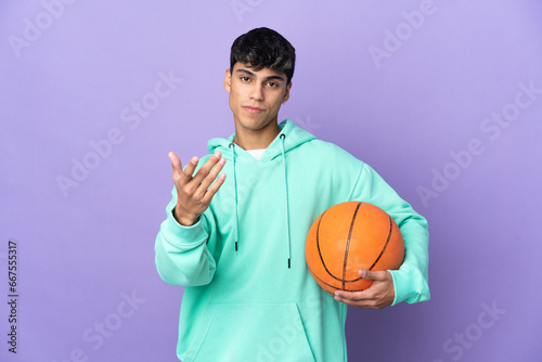 Young man playing basketball over isolated purple background inviting to come with hand. Happy that you came © luismolinero