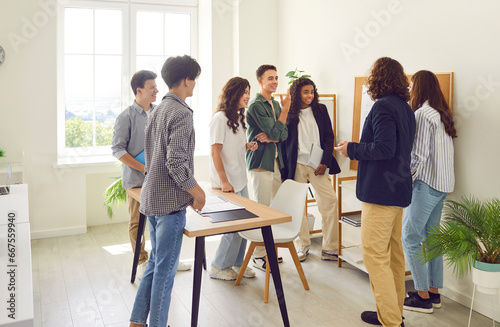 Young male teacher standing in a circle with college or high school students, talking and showing something on a board in the classroom during a lesson. Education, learning and teaching concept.