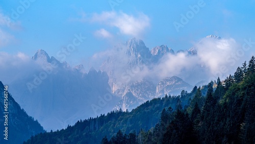 the clouds are lifting in over the mountain tops, with evergreen trees to the fore