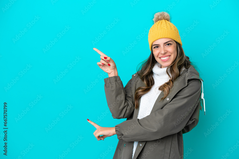 Young Italian woman wearing winter jacket and hat isolated on blue background pointing finger to the side and presenting a product Young Italian woman wearing winter jacket and hat isolated on blue background pointing finger to the side and presenting a product