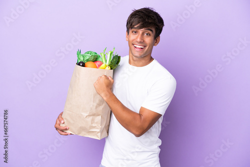 Young man holding a grocery shopping bag isolated on purple background celebrating a victory