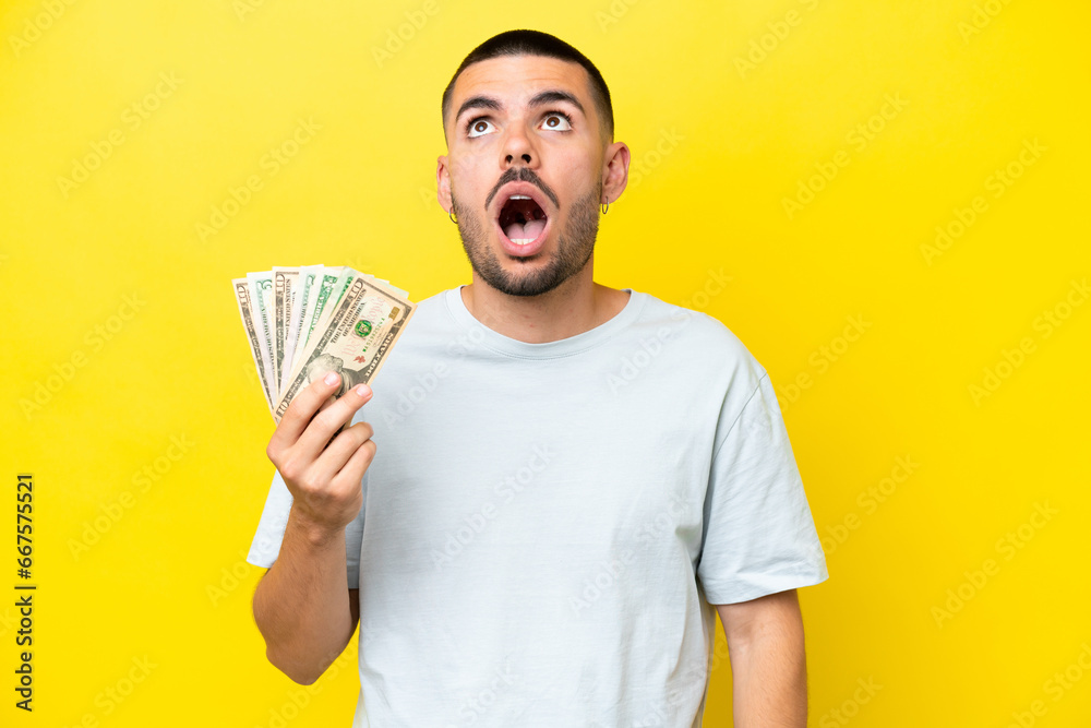 Young caucasian man taking a lot of money isolated on yellow background looking up and with surprised expression Young caucasian man taking a lot of money isolated on yellow background looking up and with surprised expression