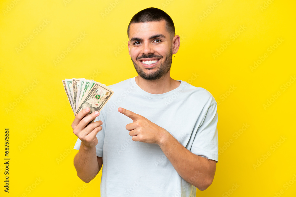 Young caucasian man taking a lot of money isolated on yellow background pointing to the side to present a product Young caucasian man taking a lot of money isolated on yellow background pointing to the side to present a product