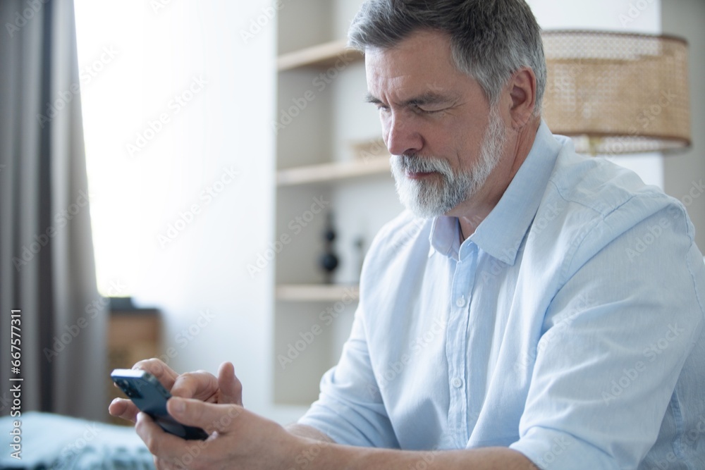 Smiling mature freelance businessman using mobile phone checking social media network or message chat sitting on chair at home. Smiling mature freelance businessman using mobile phone checking social media network or message chat sitting on chair at home.