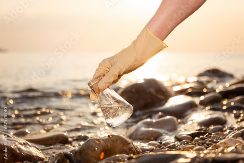 Close-up of hand in rubber glove holding glass flask with sample of pure water. In background coast and sea. Concept of Earth Day and environmental protection
