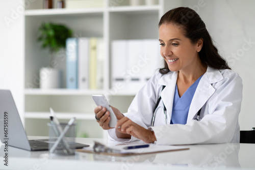 Cheerful mature woman doctor sitting in her cabinet, using gadgets