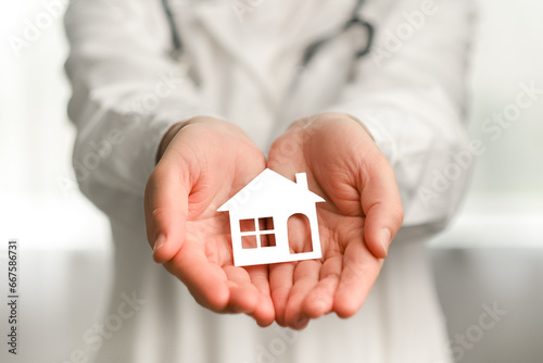 Selective focus of unknown young female doctor wearing professional medical white uniform and stethoscope holding in hands small paper house, standing in clinic. Healthcare living insurance concept