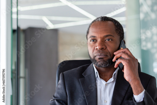 Serious confident businessman close up inside office talking on the phone, african american boss at workplace, company manager in business suit.