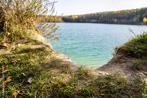 Sandy shoreline with clear turquoise lake water and ripples surrounded by dry grass and wild plants. Natural freshwater landscape background. Design for banner, poster, wallpaper. 