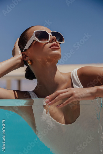 beautiful woman with dark hair in elegant swimming suit and accessories relaxing in swimming pool in summer beach club