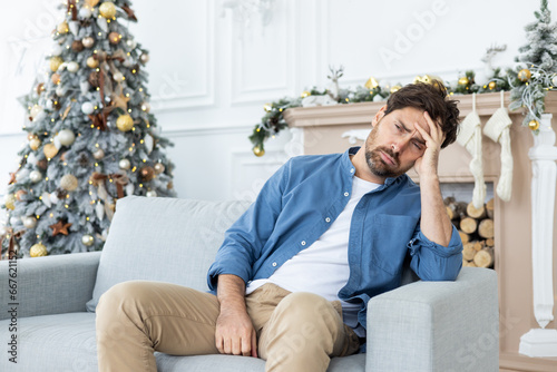 Canvastavla Tired young man sitting on sofa at home and suffering from headache and hangover after New Year's party