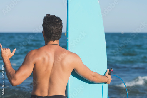 A man standing on the shore holding a surfboard - view from the back