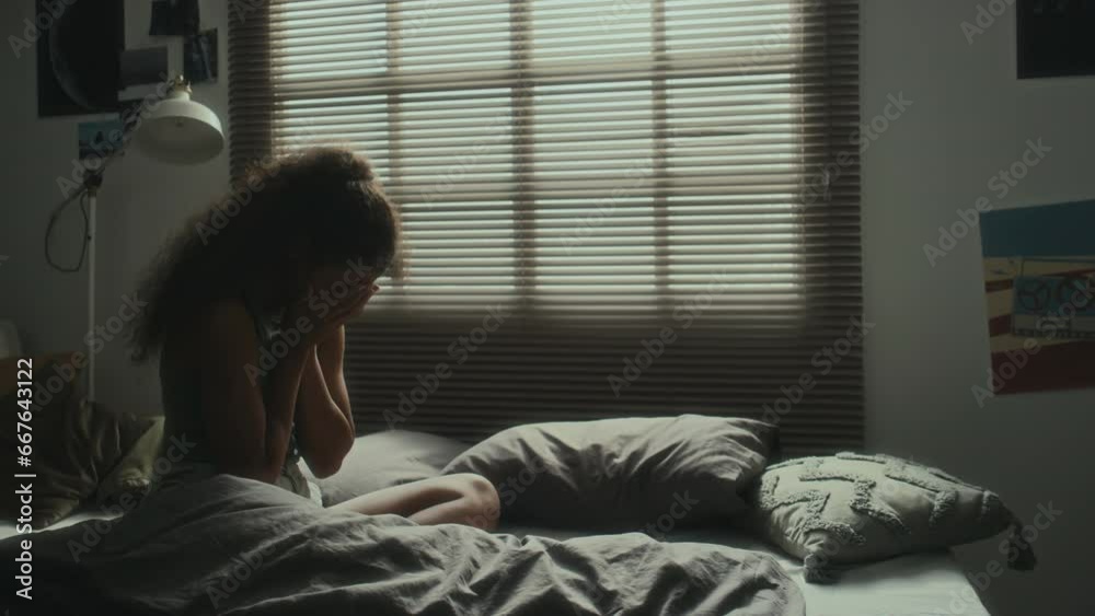 Teenage girl sitting on bed at night, reading online messages on smartphone, then getting upset and crying while suffering from cyberbullying