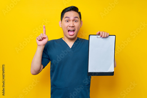 Excited professional young Asian male doctor or nurse wearing a blue uniform holding clipboard with blank paper and pointing finger up, having good ideas isolated on yellow background