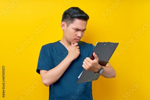 Serious professional young Asian male doctor or nurse wearing a blue uniform Hold patient diagnosis report and concentration isolated on yellow background. Healthcare medicine concept