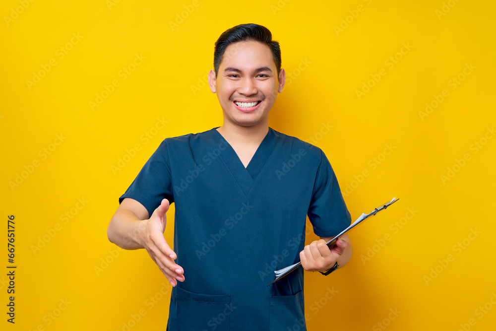 Smiling friendly professional young Asian male doctor or nurse wearing a blue uniform standing confidently holding a clipboard and greeting patients in a hospital isolated on yellow background