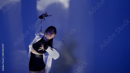 Young woman wearing a top, shorts and a shirt performing contemporary dance under the glare of light in the studio. Neon blue color scheme, shadowed background. Medium full.