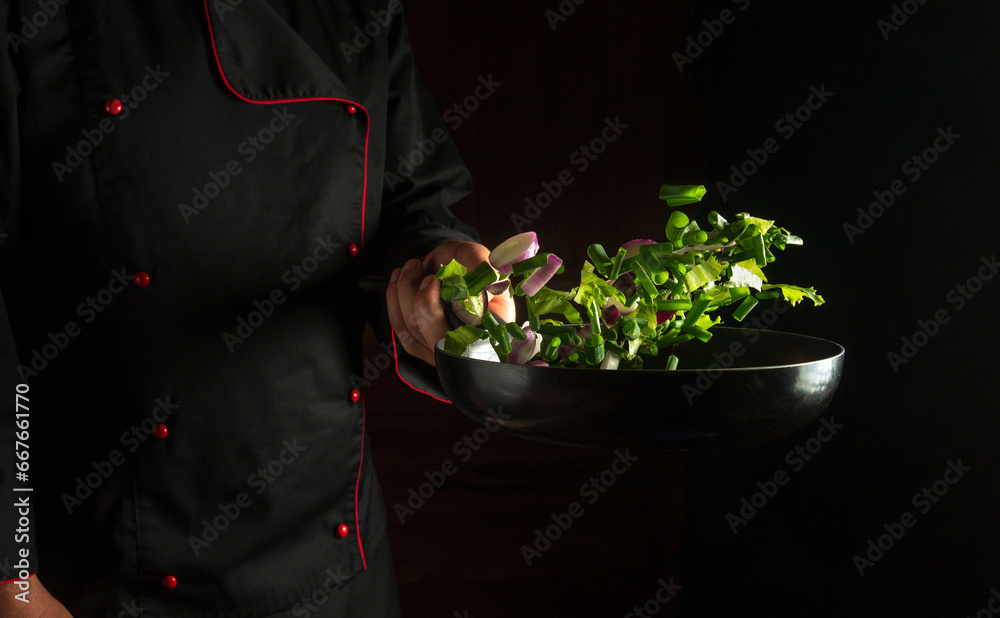The process of cooking fresh vegetables in a frying pan in the hand of a chef. Vegetable diet concept on black background