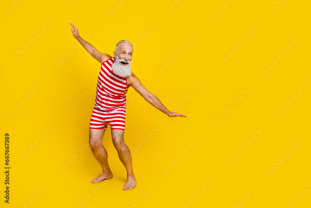 Full length photo of old man surfing ocean waves have fun at resort wearing red striped swimsuit isolated on yellow color background Full length photo of old man surfing ocean waves have fun at resort wearing red striped swimsuit isolated on yellow color background