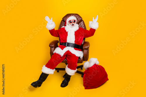 Full size portrait of eccentric impressed santa sit chair raise arms new year presents bag isolated on yellow color background