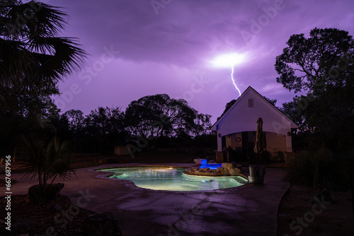 A night time lightning strike a backyard structure with a pool, a spa and trees under a purple dark cloudy sky, Hill Country, Texas