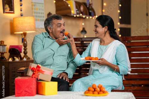 daughter and father eating sweets by sharing together at home during diwali festival celebration at home - concept of festive wishes, relationship bonding and holidays.