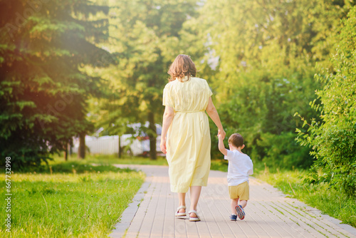 A child takes steps holding a mother woman by the hand along a path in nature. Happy baby with mom on a walk in the summer park. Kid aged about two years (one year eleven months)