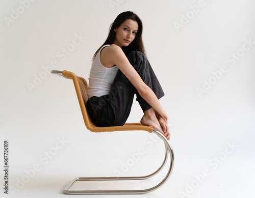 A beautiful young girl in a white top and comfortable pants sits on a chair on a white background. Casual look