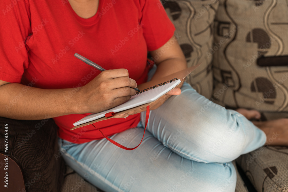 Unrecognizable Latin woman, sitting on the couch writing in a notebook, red sweater, blue jeans