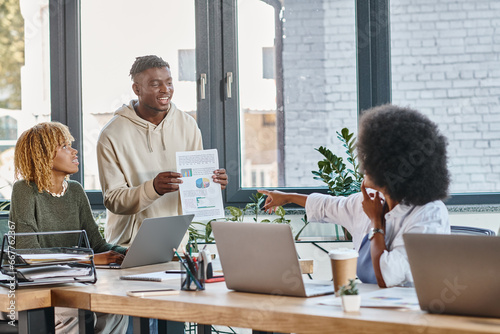 good looking african american man showing to his female friends graphics, pointing finger, coworking