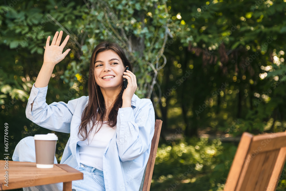 Cheerful young woman talking on the phone while sitting at the table with a cup of coffee on the background of the forest, copy space.