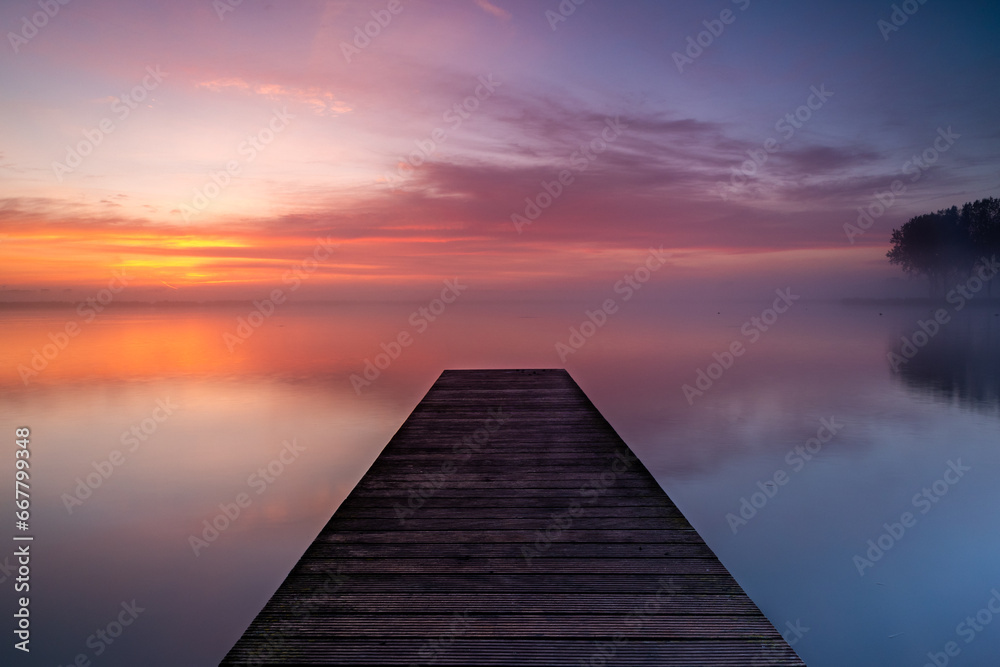 A wooden pier extends into the lake, where a layer of fog forms above the calm water. The rising sun beautifully colors the sky. - obrazy, fototapety, plakaty 