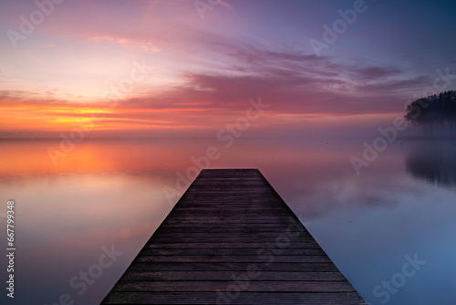 A wooden pier extends into the lake, where a layer of fog forms above the calm water. The rising sun beautifully colors the sky. photo