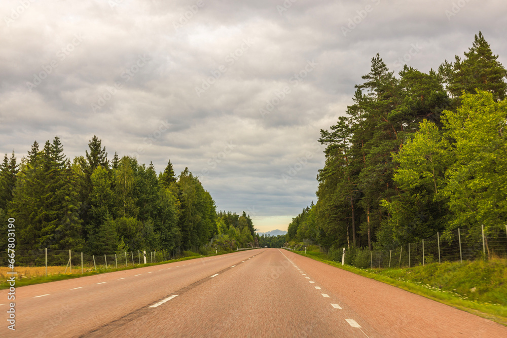 Scenic view of highway running alongside forest with trees flanking both sides and set against backdrop of cloudy sky. Sweden.