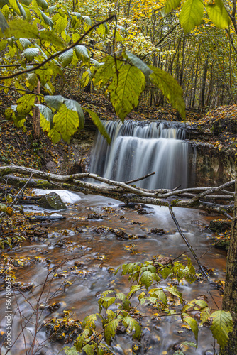 A shallow river with a waterfall flows into the canyon ravine. Water falls over two sandstone cascades. Yellow autumn leaves on land and in water. The third largest in Latvia - Dauda waterfall photo