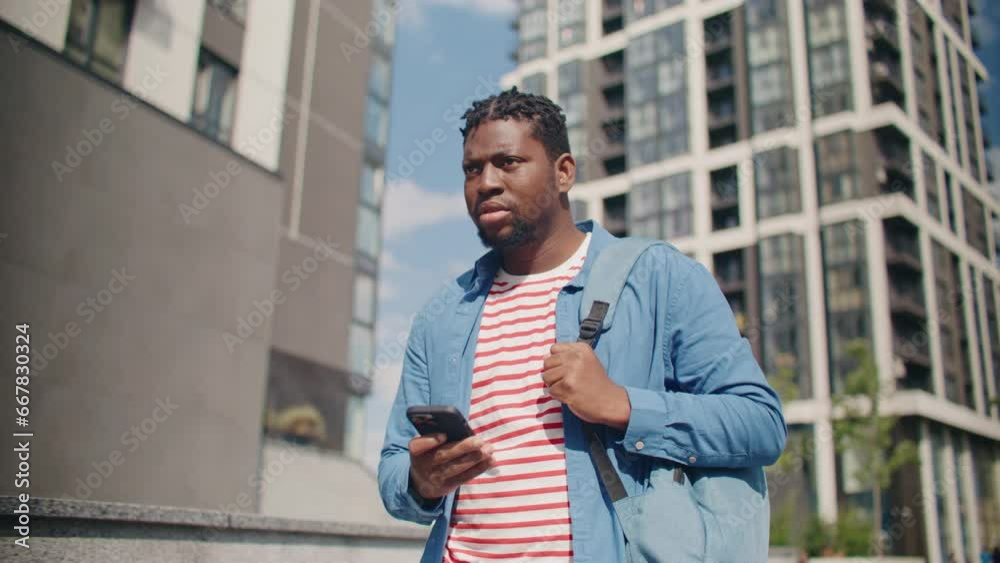 Attractive African American man carrying backpack while holding his smartphone. Actively walking down on street where beautiful woman standing. People using technology devices. Attractive African American man carrying backpack while holding his smartphone. Actively walking down on street where beautiful woman standing. People using technology devices.
