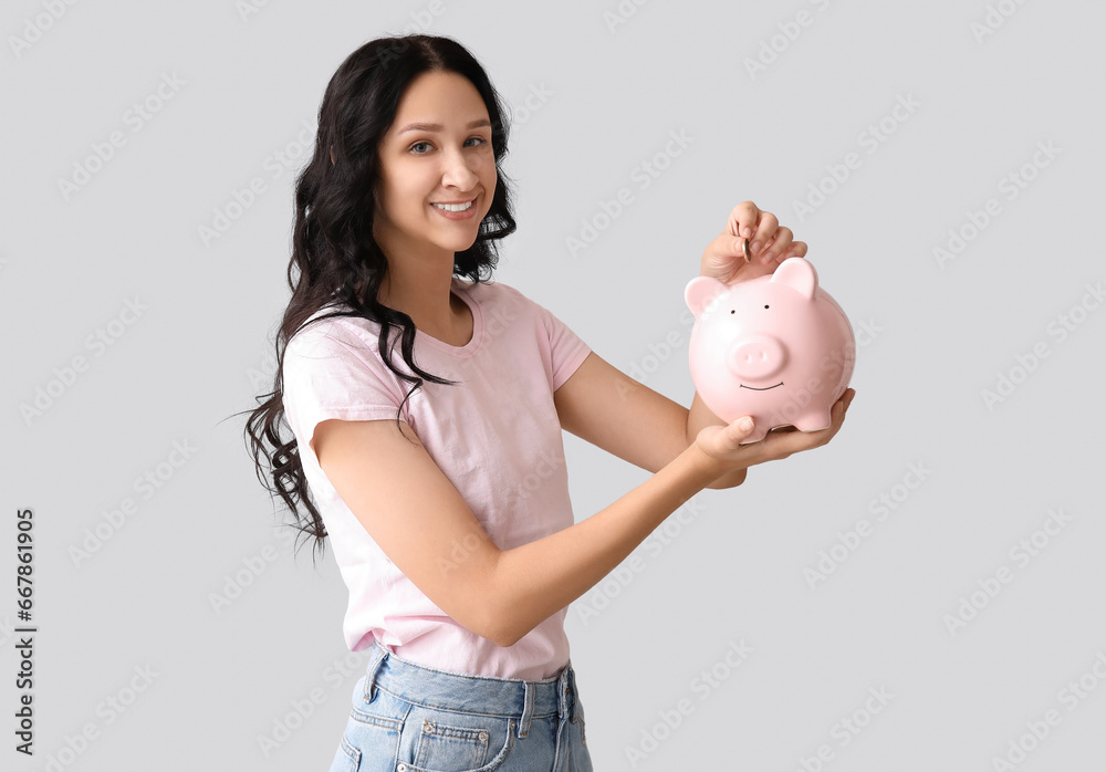 Pretty young woman putting coin into piggy bank on grey background Pretty young woman putting coin into piggy bank on grey background