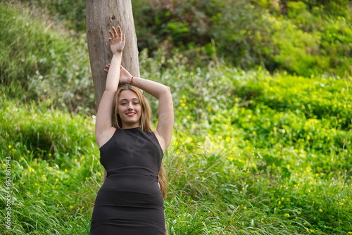 Young woman, blonde and beautiful, with a black dress, leaning on the trunk of a tree, in the middle of nature, smiling, happy and calm. Concept nature, peace, happiness, calm, trees.