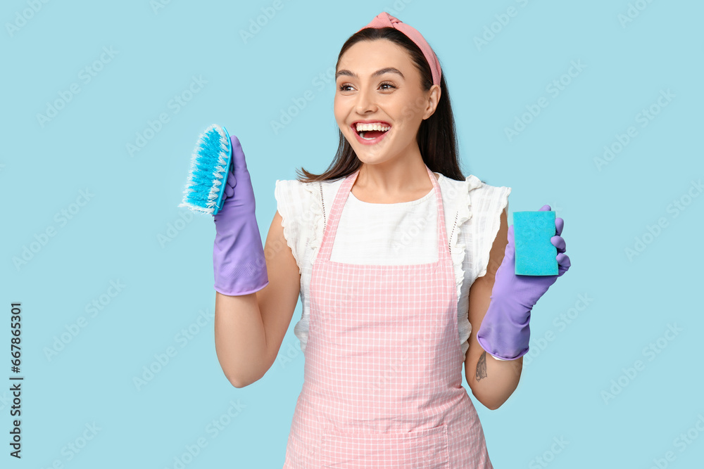 Portrait of happy young housewife in apron with cleaning brush and sponge on blue background Portrait of happy young housewife in apron with cleaning brush and sponge on blue background