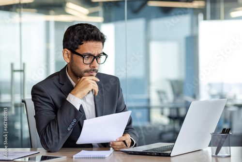 Portrait of serious pensive businessman behind paperwork, financier looking at documents, papers and contracts, thinking about solutions to set tasks, man inside office in business suit with laptop. photo