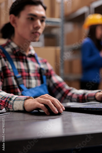 Young asian logistics manager analyzing goods supply on laptop. Warehouse worker doing inventory management on computer while sitting at desk with close up selective focus on hand