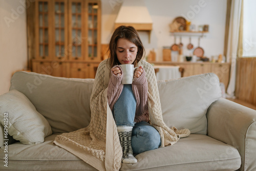 Woman freezes in winter time. Young girl wearing warm woolen socks and wrapped into plaid, holding a cup of hot drink while sitting on sofa at home. Keep warm. 