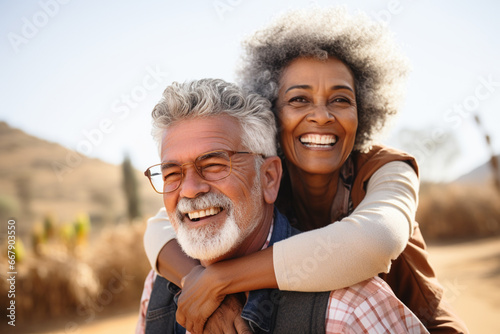 Happy mature interracial couple enjoying free time together. Active senior husband giving wife a piggyback ride while enjoying a sunny day outdoors. Energetic man and woman having fun while on holiday photo